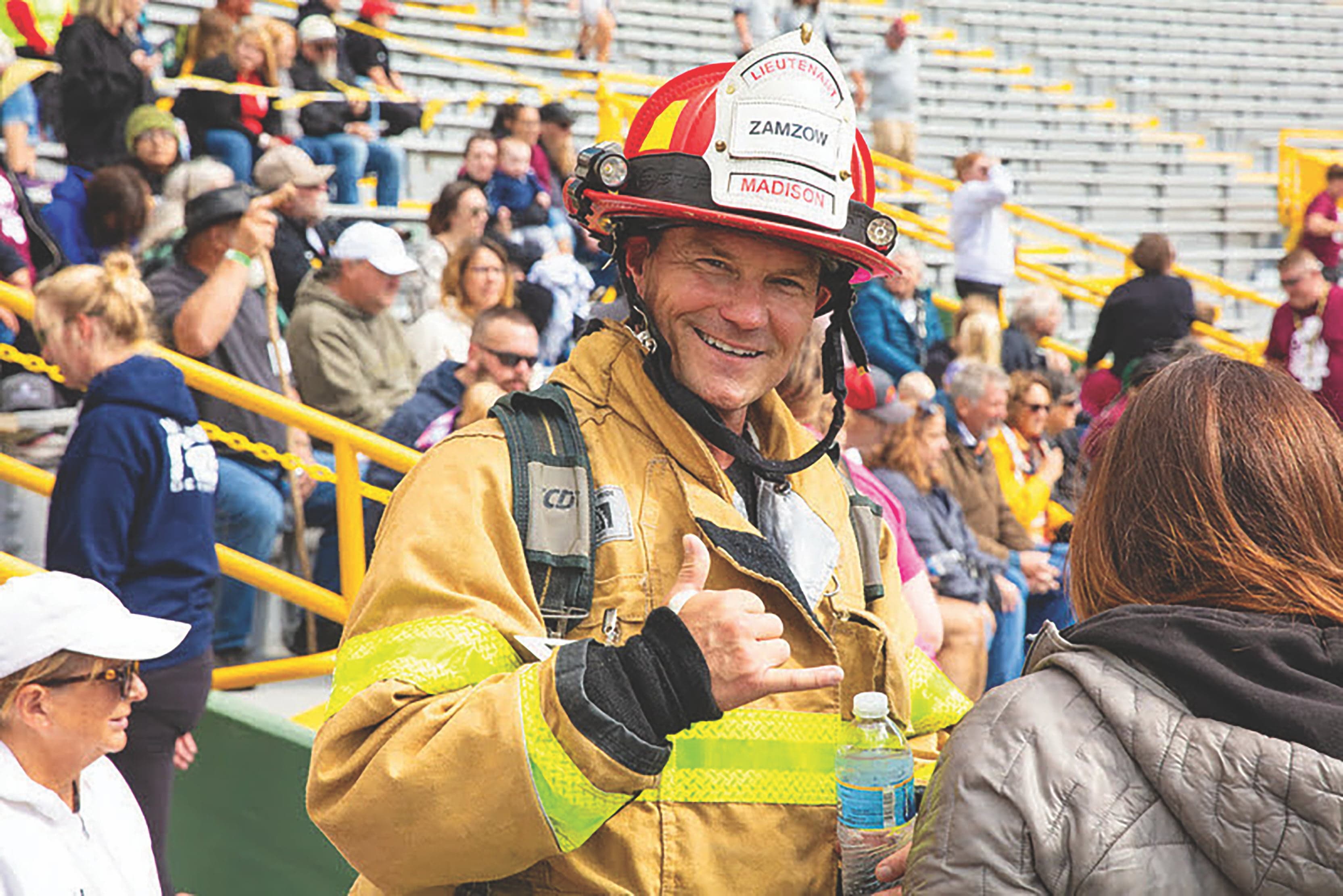 A firefighter in uniform and helmet smiles and gestures with a shaka sign while holding a water bottle. He stands in a stadium with a seated audience.