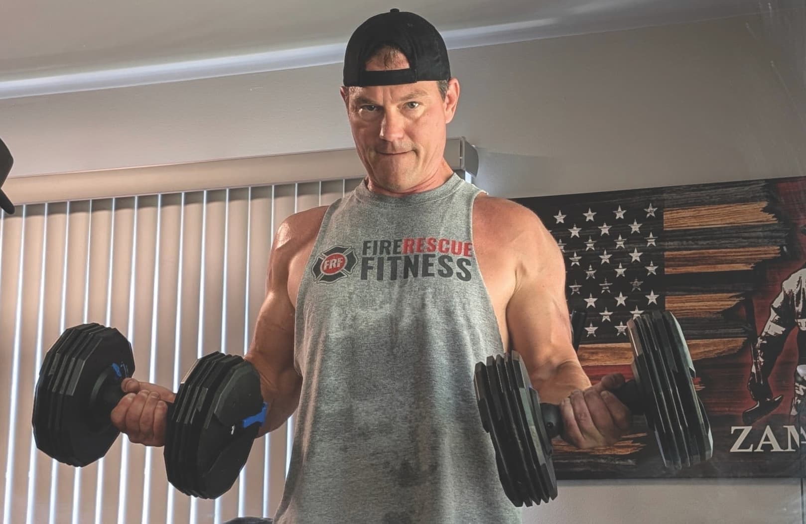 Man in a sleeveless shirt and cap lifting heavy dumbbells indoors. American flag decor and blinds in the background. Focused expression. Fitness theme.