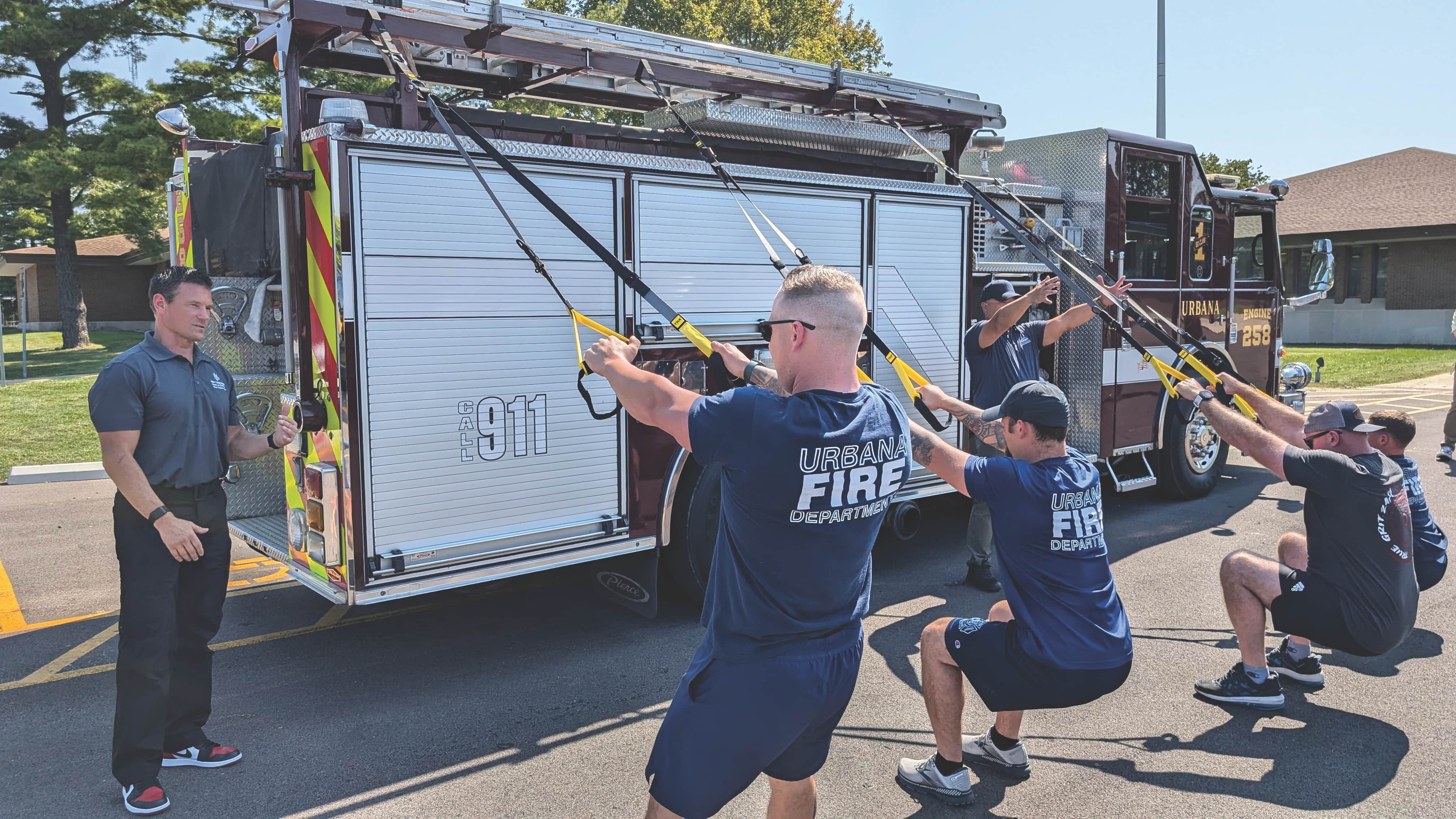 A group of firefighters perform resistance band exercises attached to a fire truck in a sunny parking lot. A trainer observes and instructs them.