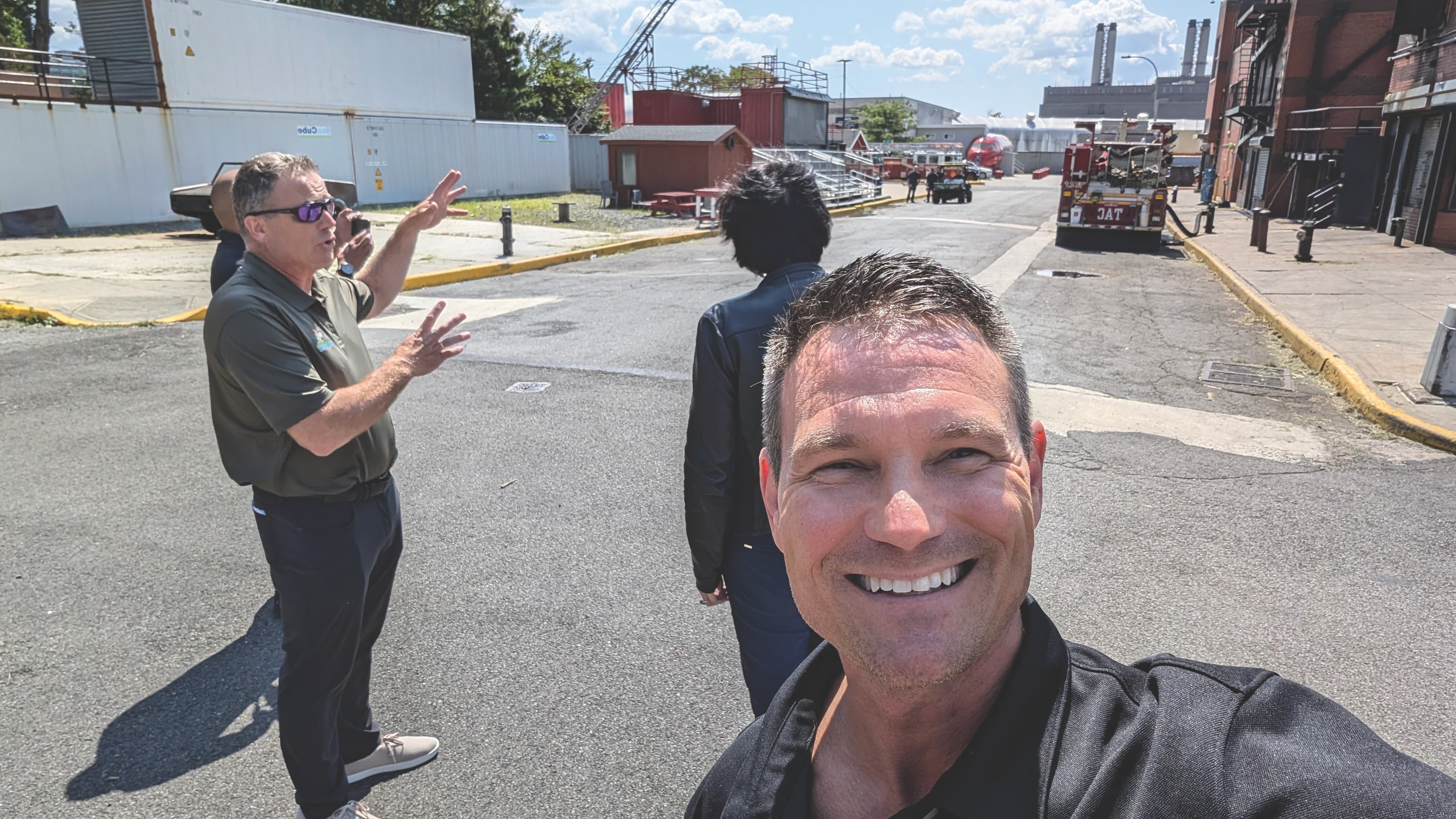 A man in the foreground takes a selfie, smiling on a sunny street. Behind him, two other people converse, one gesturing. Industrial backdrop, clear sky.